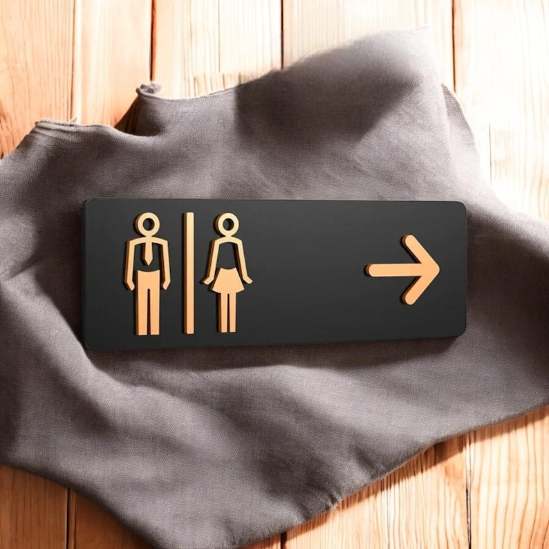 May include: A black restroom sign with gold-colored male and female figures and an arrow pointing right. The sign is on a gray cloth, with a wooden background.