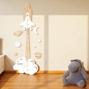 May include: A wooden height chart with a rocket ship design, measuring in inches. The chart is decorated with planets, stars, and a cloud with the name "Carter". A gray stuffed hippo sits on the floor.