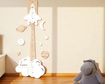 May include: A wooden height chart with a rocket ship design, measuring in inches. The chart is decorated with planets, stars, and a cloud with the name "Carter". A gray stuffed hippo sits on the floor.