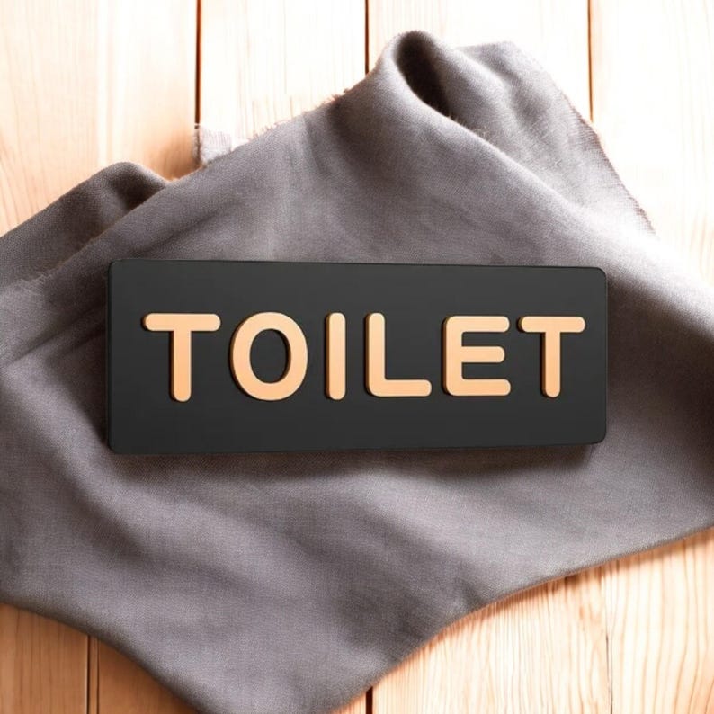 May include: A black rectangular sign with the word "TOILET" in gold lettering, resting on a gray cloth. The cloth is draped over a light brown wooden surface.