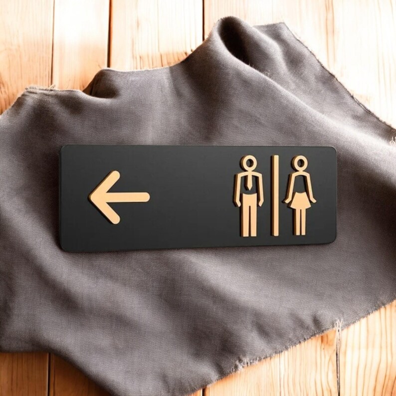 May include: A black restroom sign with a gold arrow pointing left and gold male and female figures. The sign is on a gray cloth over a wooden surface. The sign indicates the direction to the restrooms.