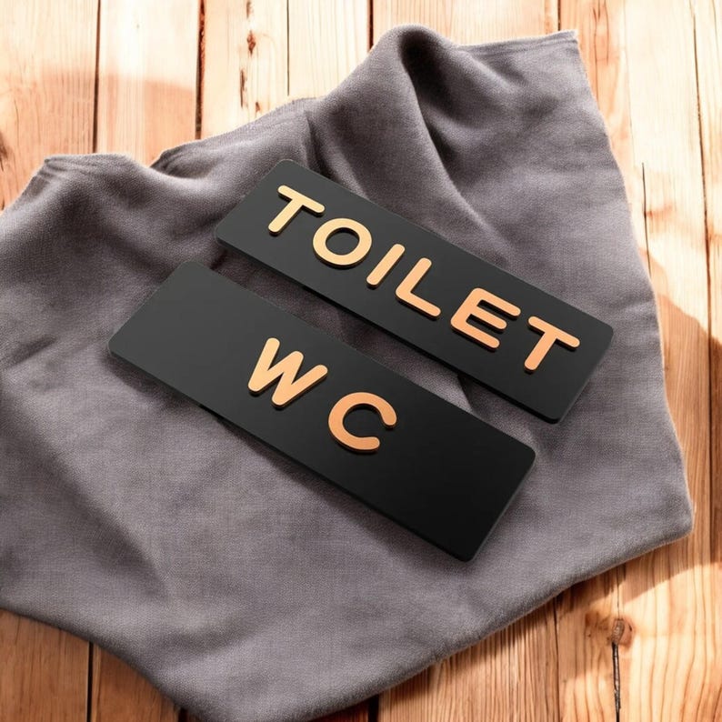 May include: Two black rectangular signs with gold lettering that read "TOILET" and "WC". The signs are resting on a gray cloth, with a wooden background. The signs are for a restroom or bathroom.