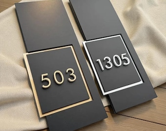 May include: Two rectangular black address plaques with numbers. One plaque has the number "503" in gold, set within a gold frame. The other has "1305" in silver, set within a silver frame. Both are on a wooden surface.