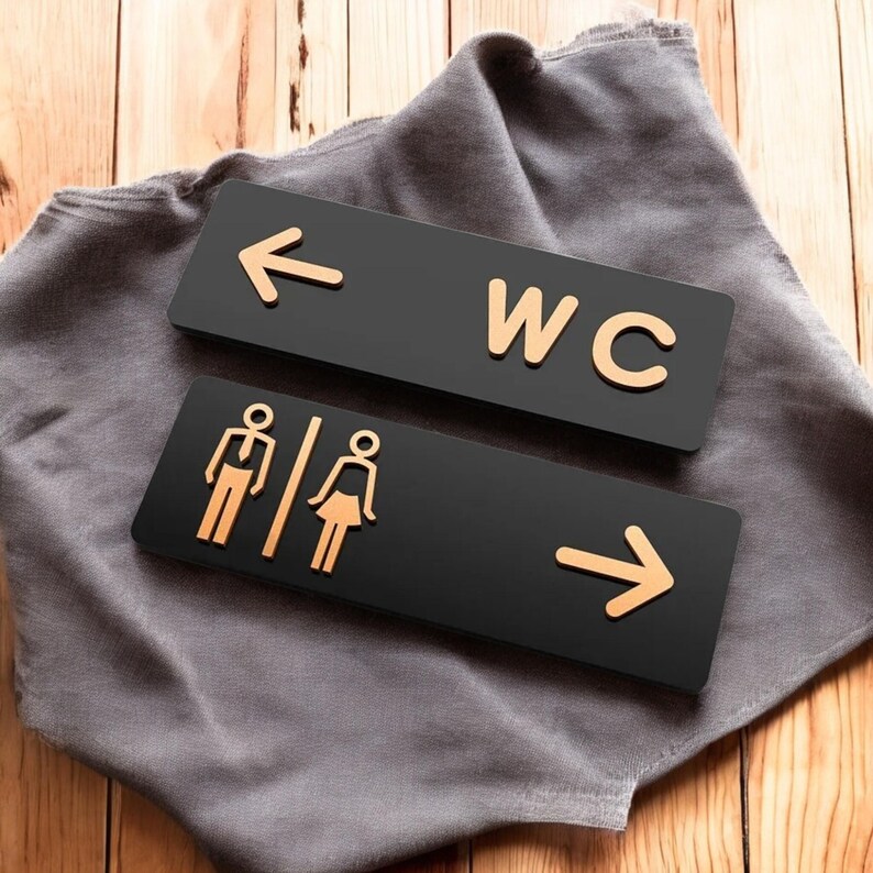 May include: Two black restroom signs with gold-colored text and symbols. One sign has the letters "WC" and an arrow pointing left. The other sign has male and female symbols and an arrow pointing right.