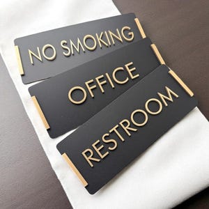 May include: Three rectangular black signs with gold lettering and accents. The signs read "NO SMOKING", "OFFICE", and "RESTROOM". The signs are likely for a business or public space.