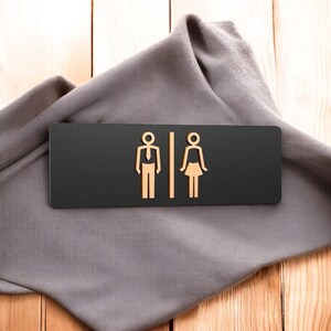 May include: A black rectangular restroom sign with gold-colored male and female figures. The sign is resting on a gray cloth, with a wooden background.