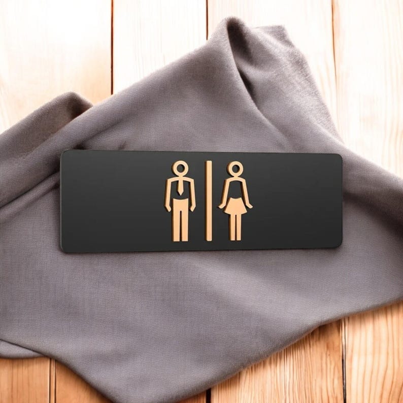 May include: A black rectangular restroom sign with gold-colored male and female figures. The sign is resting on a gray cloth, with a wooden background.