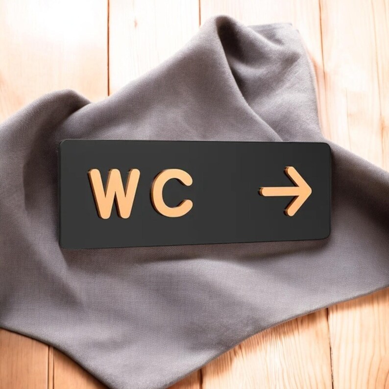 May include: A black rectangular sign with the letters "WC" in gold and an arrow pointing right. The sign is on a gray cloth, set against a light wood background.