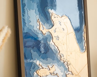 May include: A framed 3D bathymetric map featuring a light wood landmass with engraved roads and a compass rose, set against layered shades of blue resin representing varying ocean depths, all enclosed within a dark brown rectangular frame.