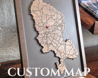 May include: A framed wooden map cutout featuring engraved road networks and a small red heart marker placed over the location labeled Offemont. The map is mounted on a dark gray background within a dark brown wooden frame, displayed on a stand.