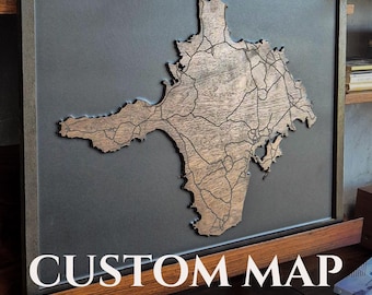 May include: A rectangular framed wall art piece featuring a wooden cutout of a landmass with engraved road lines, mounted on a solid dark gray background. The text CUSTOM MAP is printed in white capital letters at the bottom of the frame.