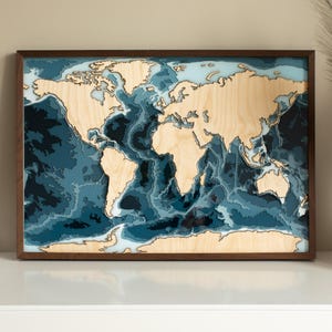 May include: A framed wooden world map with a dark brown frame. The continents are light-coloured wood, and the oceans are depicted in shades of blue. The map is displayed on a white surface with a vase and books in the background.