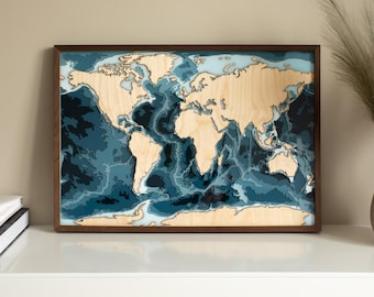 May include: A rectangular framed wall art piece featuring a layered wooden world map. The landmasses are depicted in a light natural wood tone, while the ocean depths are represented by various shades of blue, ranging from light teal to deep navy.
