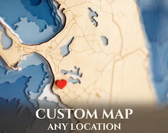 May include: A layered wooden bathymetric map featuring light tan landmasses with engraved roads and deep blue water depths. A small red heart-shaped marker is placed on the land. The text CUSTOM MAP ANY LOCATION is printed at the bottom.