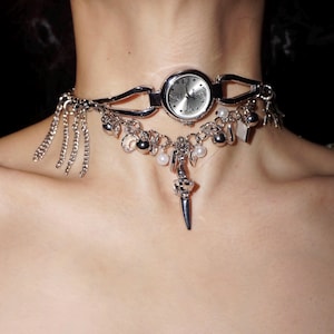 Puede incluir: Un collar choker plateado con un reloj, abalorios y un colgante que cuelga. El collar presenta una variedad de abalorios, incluyendo cuentas, perlas y un pequeño colgante con forma de espada.