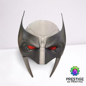 May include: A 3D printed black and gray mask with red eyes. The mask has a textured surface and is shaped like a superhero's mask.