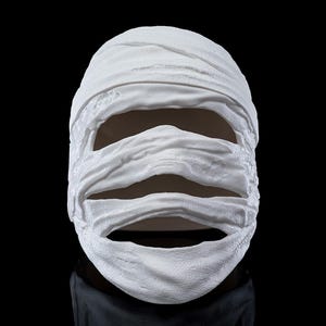 Puede incluir: Máscara de momia blanca con tres agujeros para los ojos y un agujero para la boca. La máscara está hecha de un material texturizado que se asemeja a vendas.