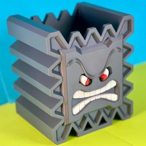 May include: A gray 3D printed pencil holder shaped like a cartoon face with a frowning expression. The face has red eyes and white teeth.