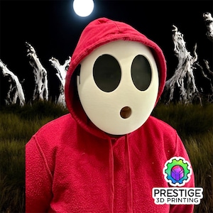 Peut inclure: Une personne portant un sweat à capuche rouge et un masque blanc de Shy Guy avec de grands trous noirs pour les yeux et une petite ouverture pour la bouche. L'arrière-plan présente une scène de nuit sombre avec une pleine lune et des arbres en silhouette. Le logo de Prestige 3D Printing est visible.