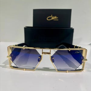 May include: Gold-framed sunglasses with blue gradient lenses. The geometric lenses are set in a gold frame with a black top bar. A black box with gold lettering is in the background.