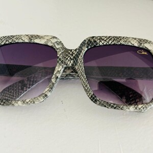May include: Gray and black square sunglasses with a snakeskin pattern and gold accents. The sunglasses have a dark purple lens and the brand name 