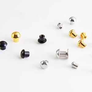 Op de afbeelding: Een assortiment metalen klinknagels in verschillende kleuren, waaronder goud, zilver en zwart, liggen op een witte ondergrond. De klinknagels hebben een afgeronde, paddenstoelvormige kop en een pen voor bevestiging, geschikt voor leerbewerking of kledingconstructie.