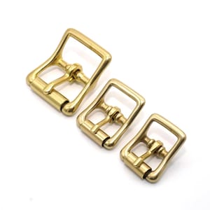 May include: Three brass roller buckles of varying sizes are arranged diagonally on a white background. Each buckle has a rectangular frame with a roller and a tongue for securing straps. The buckles have a shiny, golden finish.