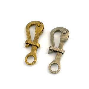 Könnte beinhalten: Zwei Metall-Federkarabinerhaken. Einer ist messingfarben, der andere silberfarben. Jeder Haken hat eine gebogene Oberseite, einen federbelasteten Riegel und einen Ring am Boden. Die Haken sind zum Sichern von Gegenständen bestimmt.