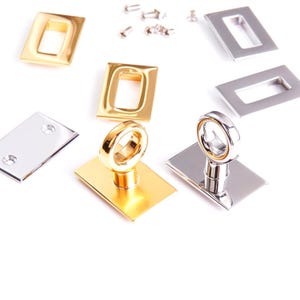 May include: An assortment of gold and silver-toned metal hardware pieces. The hardware includes rectangular frames, rectangular plates, and ring-shaped pulls. Small screws are scattered across the top. The pieces are displayed on a white background.