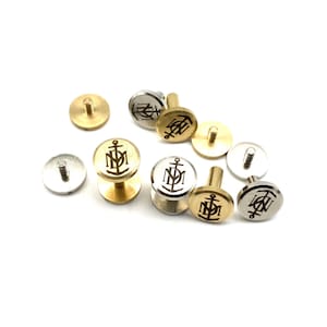 May include: A collection of gold and silver metal buttons with an anchor and initials engraved on the top. The buttons have a screw-on back.