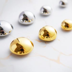 Peut inclure: Un assortiment d'objets décoratifs métalliques brillants. Certains sont argentés, d'autres dorés, et tous ont une forme lisse et arrondie. Les objets sont dispersés sur une surface blanche avec de subtiles veines, créant une esthétique moderne et élégante.