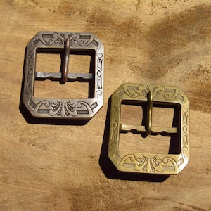 Op de afbeelding: Twee decoratieve metalen gespen. Eén is zilverkleurig, de andere is messingkleurig. Beide hebben een achthoekige vorm met sierlijke krullen en een centrale staaf voor de riem. De gespen worden op een houten oppervlak getoond.