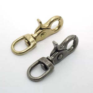 Könnte beinhalten: Zwei Metall-Drehhaken. Einer ist goldfarben glänzend, der andere dunkelgrau matt. Beide haben einen federbelasteten Hebel und einen D-Ring zum Befestigen. Die Verschlüsse befinden sich auf weißem Hintergrund.