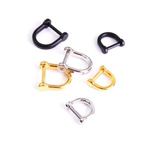 May include: An assortment of D-shaped shackles in various metallic finishes, including black, gold, and silver. The shackles are arranged on a white background, showcasing their design and construction. These hardware components are used for connecting ropes, chains, and other rigging.