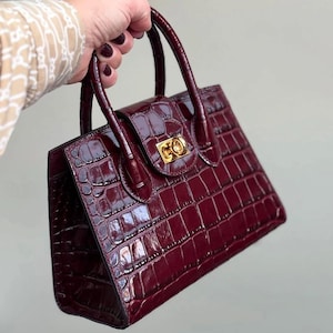 Pu&ograve; includere: Una borsa bordeaux con una texture a pelle di coccodrillo. La borsa ha una forma rettangolare, un manico superiore e una chiusura dorata. La borsa &egrave; tenuta da una persona.