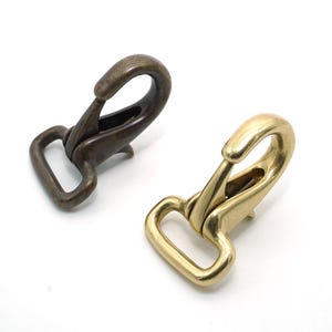 Könnte beinhalten: Zwei Metall-Drehhaken. Einer ist dunkelbronze, der andere glänzend goldfarben. Die Haken haben einen federbelasteten Verschluss für sicheren Halt. Sie werden oft beim Basteln und bei Lederarbeiten verwendet.