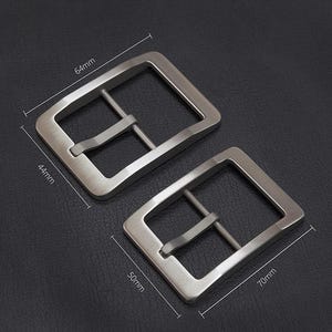Könnte beinhalten: Zwei rechteckige Gürtelschnallen in Silber. Die größere Schnalle misst 64 mm x 44 mm, die kleinere 70 mm x 50 mm. Jede Schnalle hat eine Mittelstange für den Gürtelriemen. Die Schnallen werden auf dunklem Untergrund gezeigt.