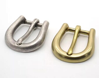 Könnte beinhalten: Zwei Metallschnallen. Eine ist silberfarben, die andere goldfarben. Beide haben eine klassische U-Form mit einem Mittelsteg für den Gürtelriemen. Die Schnallen werden vor weißem Hintergrund gezeigt.
