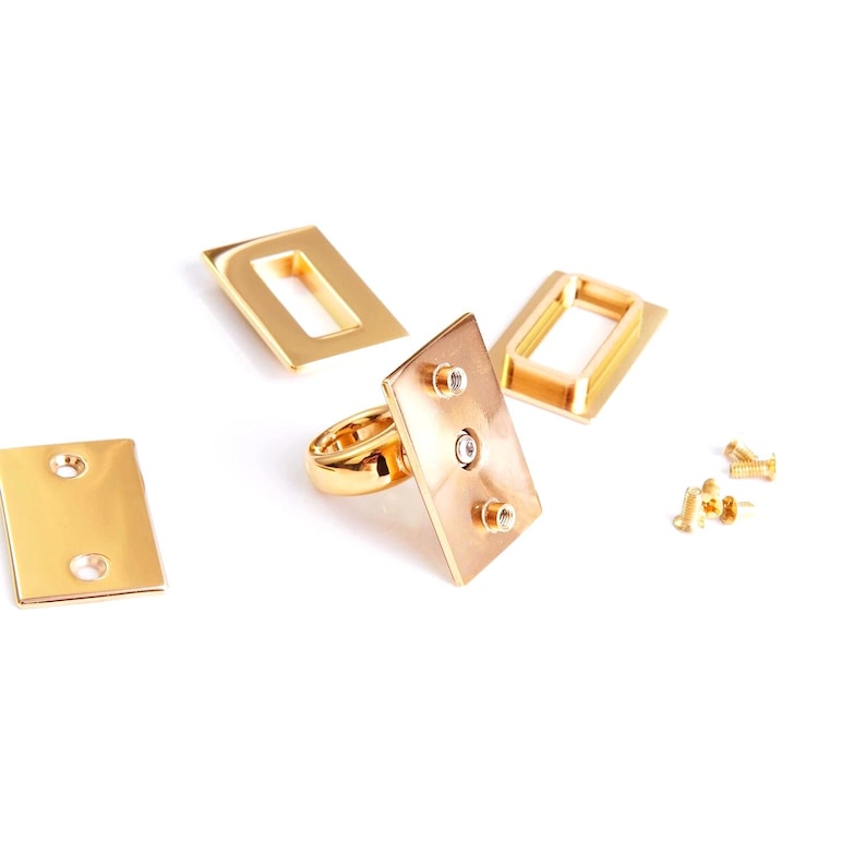 Pu&ograve; includere: Una collezione di componenti di gioielli in metallo color oro, tra cui un anello con una faccia rettangolare e piccoli abbellimenti rotondi. Ulteriori pezzi rettangolari e quadrati sono sparsi.