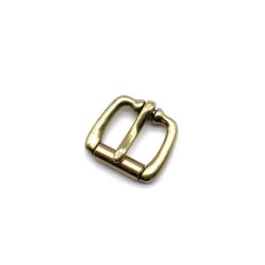 Pode incluir: Uma fivela de metal dourado com formato quadrado e acabamento liso. A fivela tem um design simples e é adequada para uma variedade de artigos de couro.