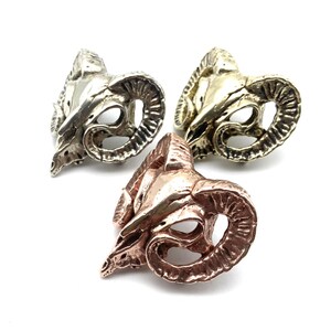 May include: Three ram skull sculptures in different metal finishes: silver, gold, and copper. The sculptures are detailed and realistic, with intricate horns and facial features.