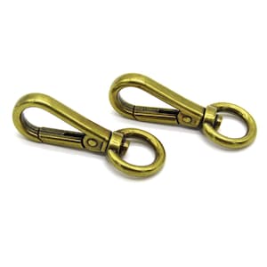May include: Two antique brass-colored swivel snap hooks. Each hook has a spring-loaded clasp and a circular ring at the base. The hooks are designed for attaching to bags, straps, or other items.