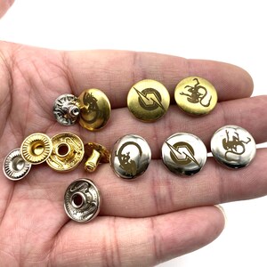 Op de afbeelding: Een verzameling gouden en zilveren metalen knopen met verschillende gegraveerde ontwerpen. De ontwerpen omvatten een bliksem, een draak en een gestileerde letter "S".