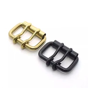 May include: Two metal roller buckles. One is gold-toned, the other is black. Each buckle has a rectangular frame with a central roller bar and two bars for the belt strap. The buckles are displayed on a white background.