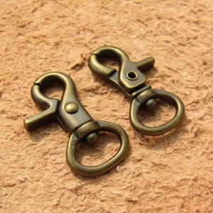 Puede incluir: Dos mosquetones giratorios de color latón antiguo. Cada gancho tiene un cierre de resorte y un anillo circular. Los ganchos se muestran sobre una superficie texturizada de color marrón claro.