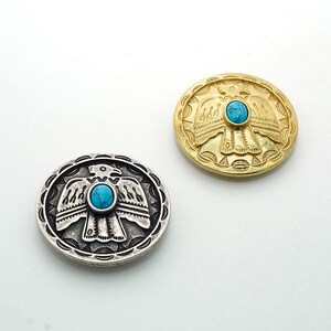 以下が含まれることがあります： 鷲のデザインと中央にターコイズの石が付いた、2つの丸い金属製のボタン。1つのボタンはシルバー色、もう1つは金色です。ボタンは、縁に詳細なエンボス加工のデザインが施されています。