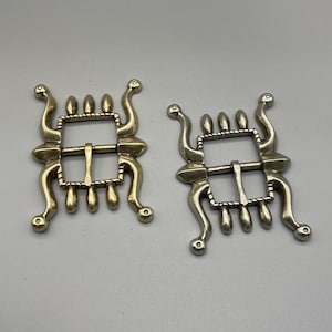 May include: Two antique metal buckles with an ornate design. Each buckle has a rectangular frame with a central bar and a series of decorative elements, including curved arms and teardrop shapes. The buckles are a muted gold color.