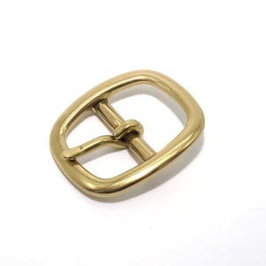 Puede incluir: Primer plano de una hebilla de cinturón de latón. La hebilla tiene forma rectangular con esquinas redondeadas y una barra central. El metal tiene un acabado dorado brillante, sobre fondo blanco.