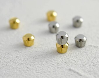 May include: A collection of small, metallic decorative objects. Some are gold-colored, while others are silver. The objects have a rounded, fluted base and a flat top. They are scattered on a textured, white surface.