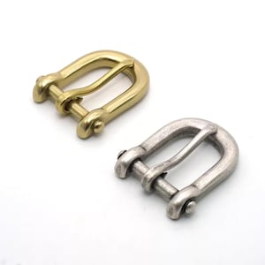 Puede incluir: Dos hebillas de arnés metálicas. Una es de color dorado brillante y la otra de color plateado mate. Ambas hebillas tienen forma de U con una barra central y extremos redondeados. Probablemente se utilizan para marroquinería o proyectos similares.